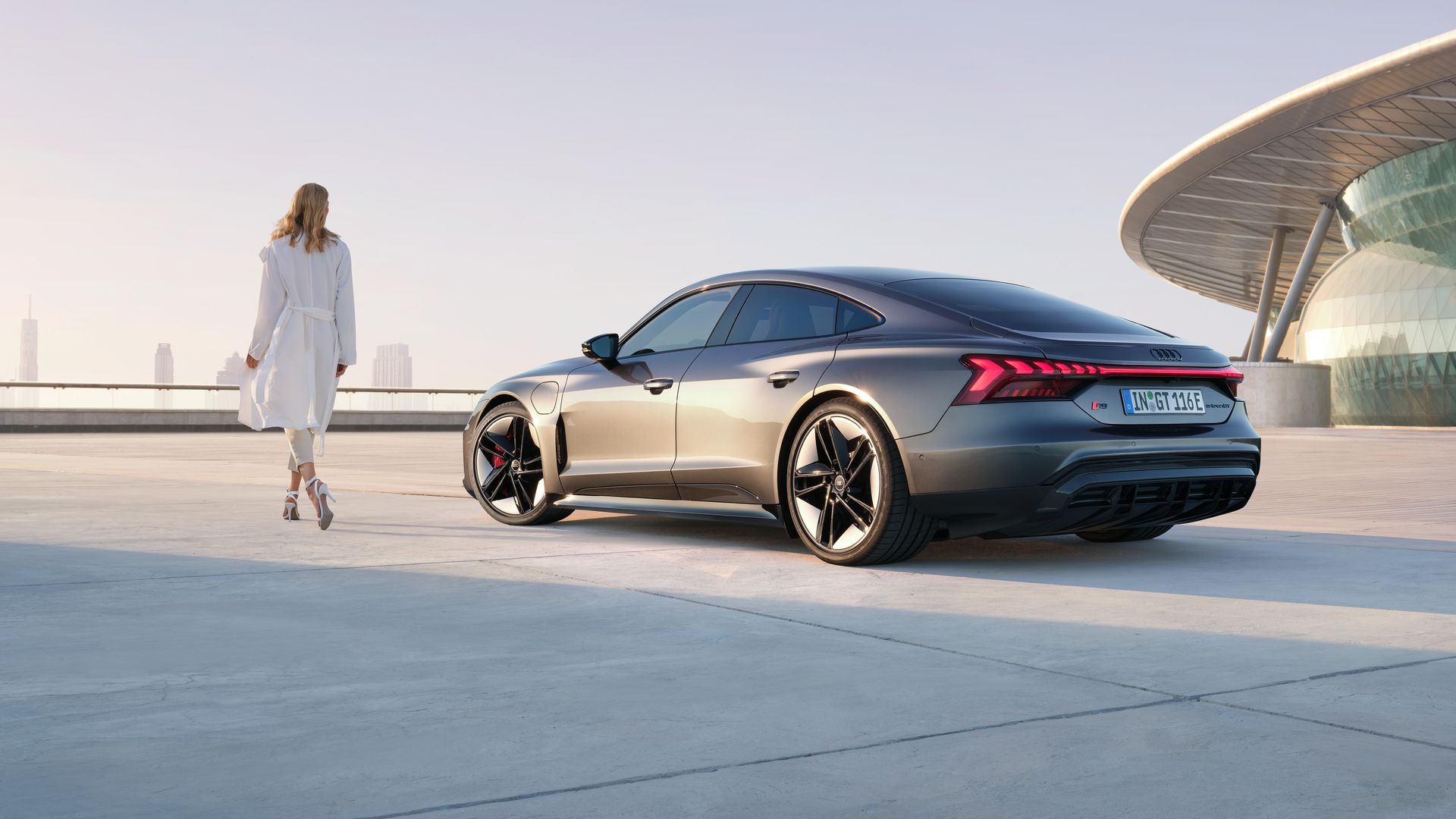 Elegante Frau neben Sportwagen Audi RS e-tron GT auf einer Betonfläche vor futuristischer Architektur.