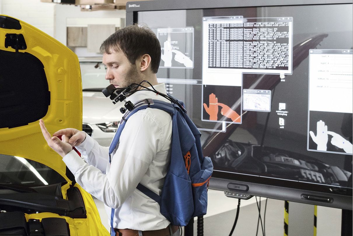 Das Foto zeigt einen Audi Mitarbeiter mit einem Rucksack auf dem Rücken, der eine technische Vorrichtung zur Hand-Projektion enthält. Der Techniker erprobt diese Projektionstechnologie und blickt dabei auf seine Hand. 