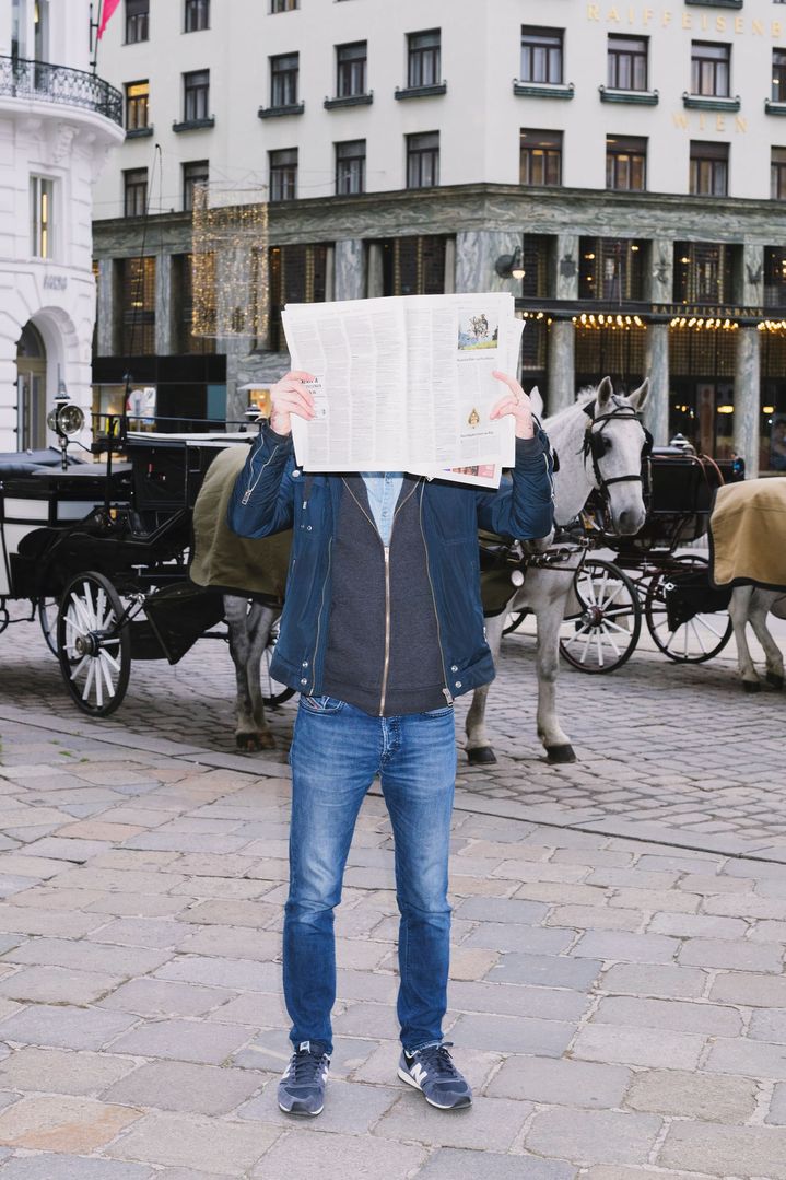 Daniel Cronin stands on Vienna’s Michaelerplatz square near the Spanish Riding School. An open newspaper obscures his face. Horse-drawn carriages with tourists are passing behind him.