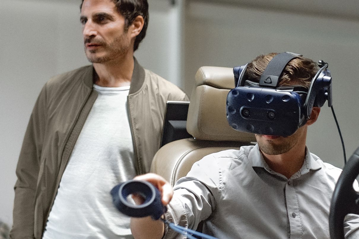 Gael Buzyn steht hinter einer Person, die mit VR-Brille in einem Fahrzeugsitz sitzt.
