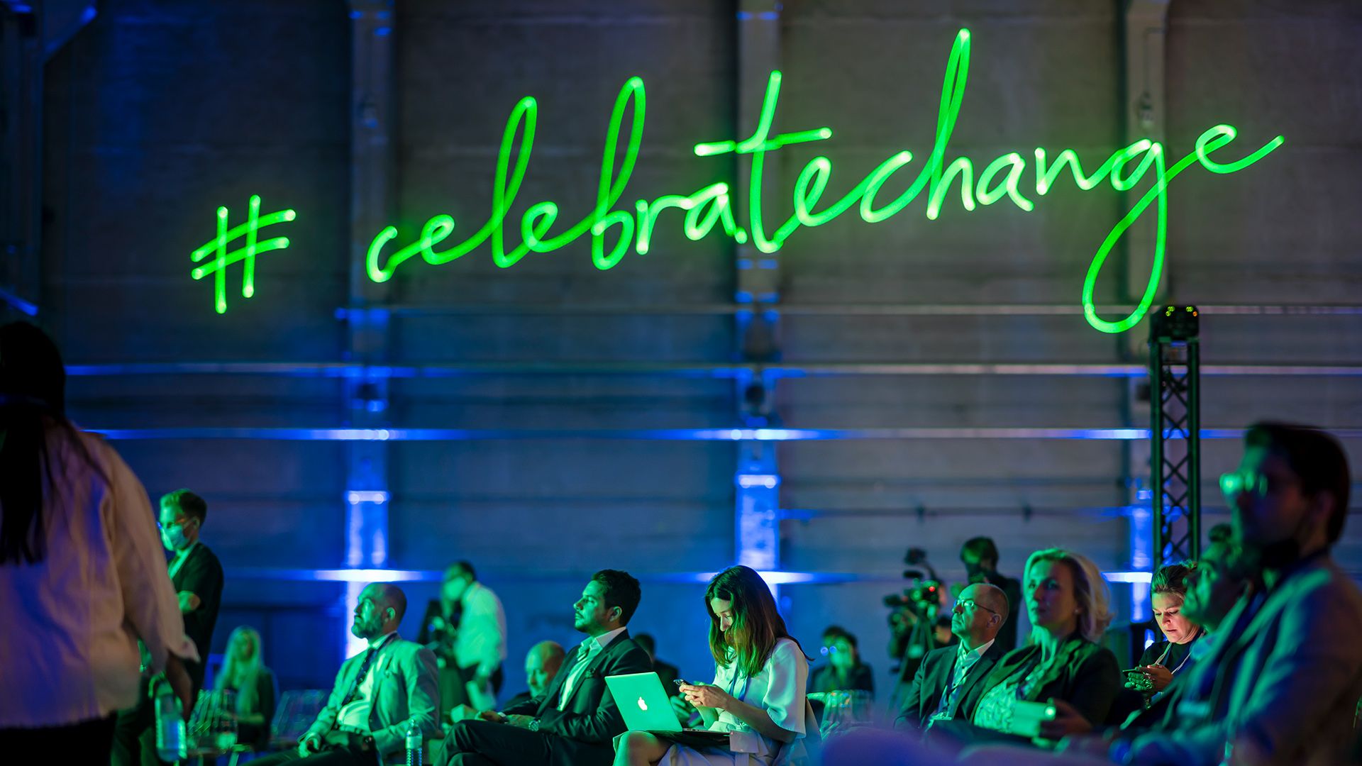 De groene tekst „#celebratechange“ geeft licht boven de hoofden van meerdere mensen in het publiek. 