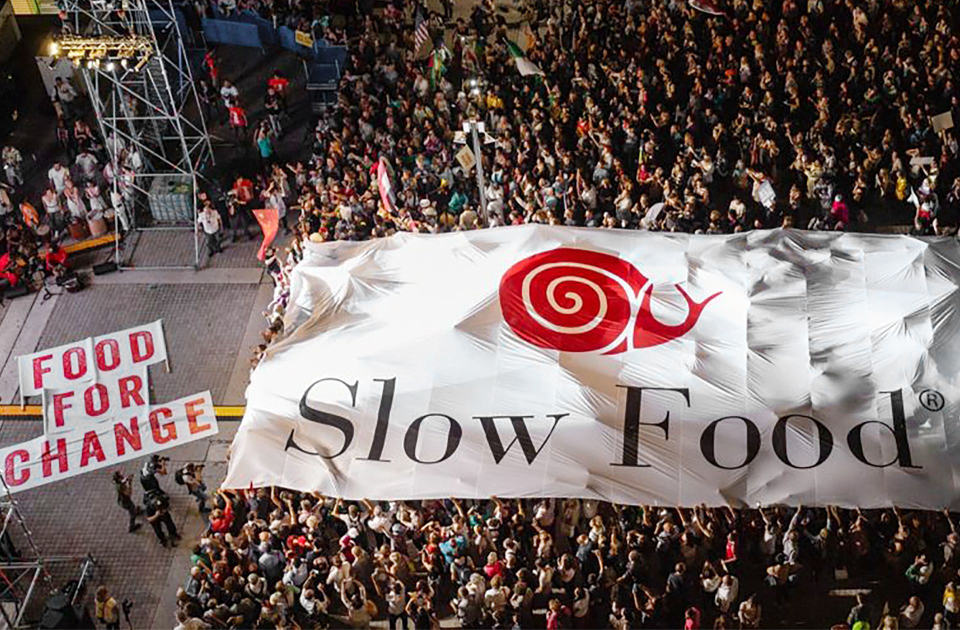 Büyük bir Slow Food pankartı taşıyan kalabalık bir grup, bir sahnenin önünde duruyor.