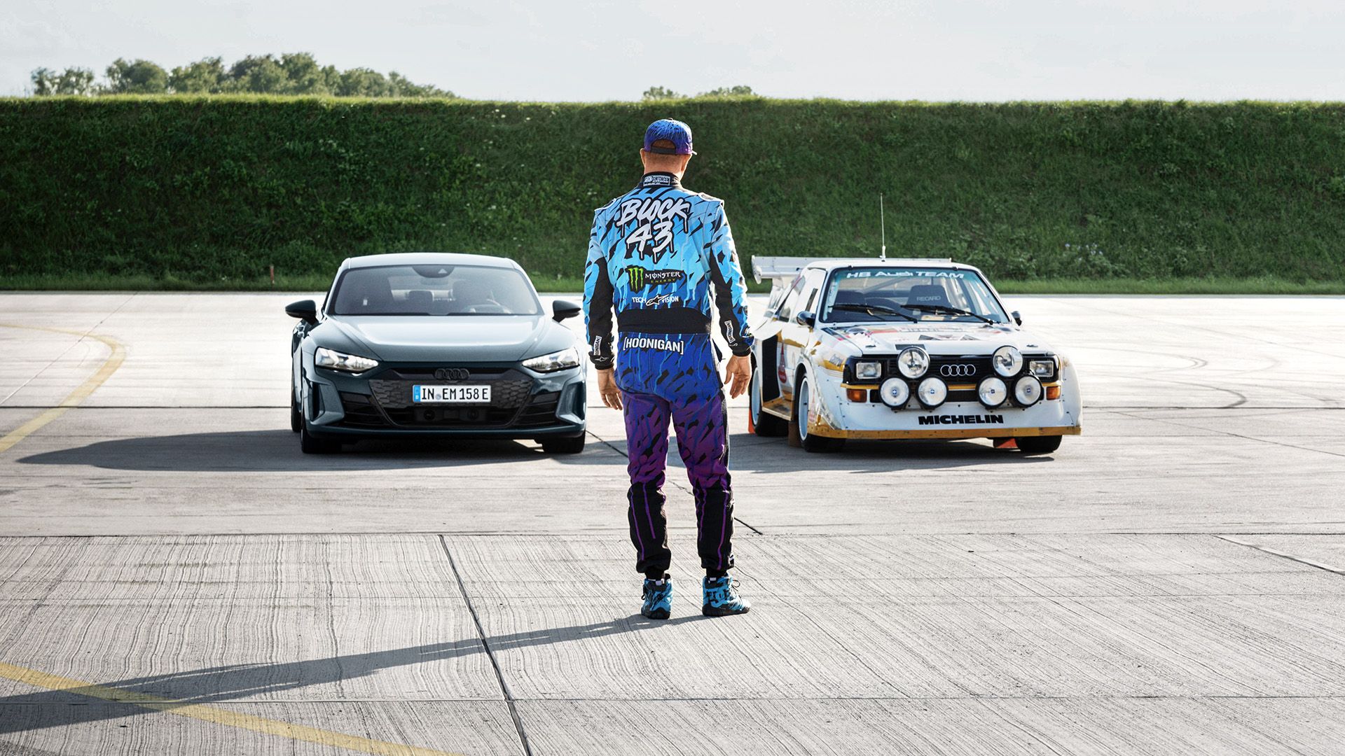 Ken Block stands with his back to the camera in front of two Audi models.
