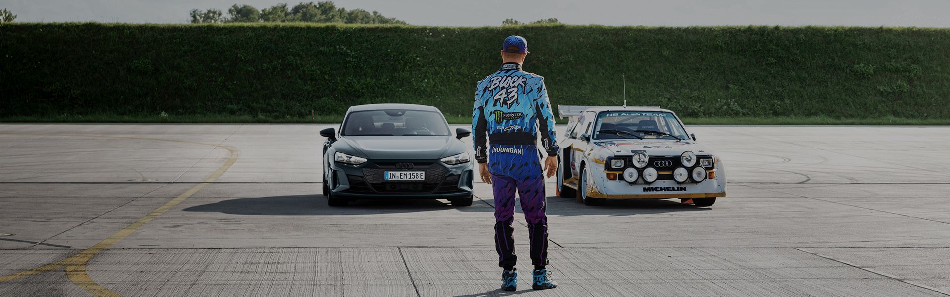 Ken Block stands with his back to the camera in front of two Audi models.