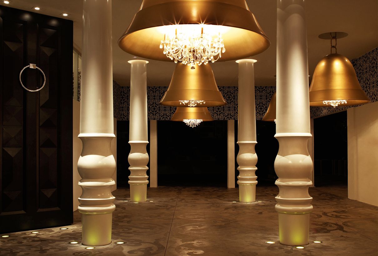Bir salonda beyaz sütunlar arasında asılı, çan şeklindeki altın lambalar.
