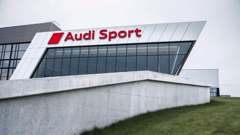 Detrás de un pequeño muro se ve el edificio de Audi Sport.