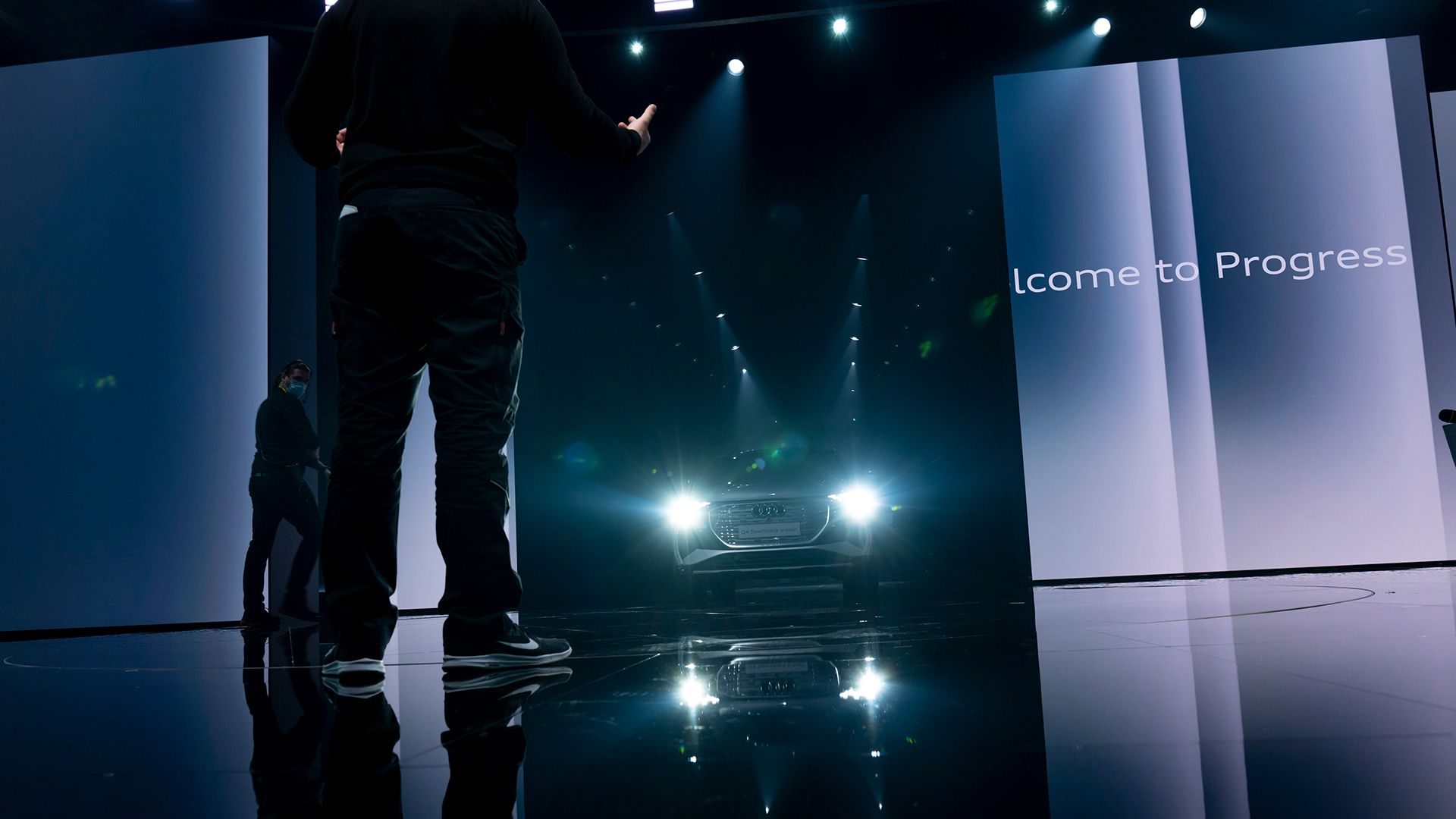Der Audi Q4 e-tron fährt auf die Bühne, die Scheinwerfer leuchten. Rechts ist ein Bildschirm mit dem Schriftzug „Welcome to Progress“.