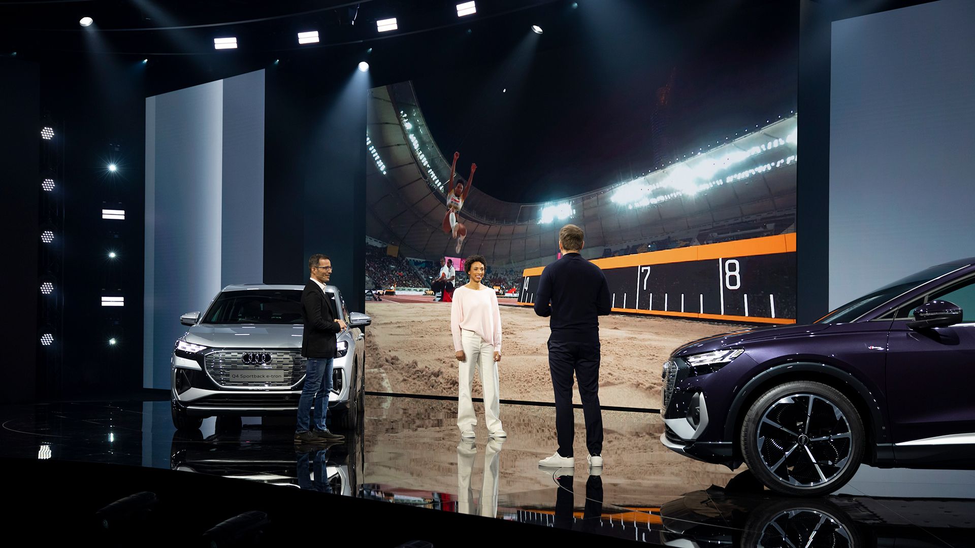 Malaika Mihambo, Steven Gätjen en Marcus Keith staan op het toneel voor een scherm, waarop de sportvrouw net uit de lucht springt.