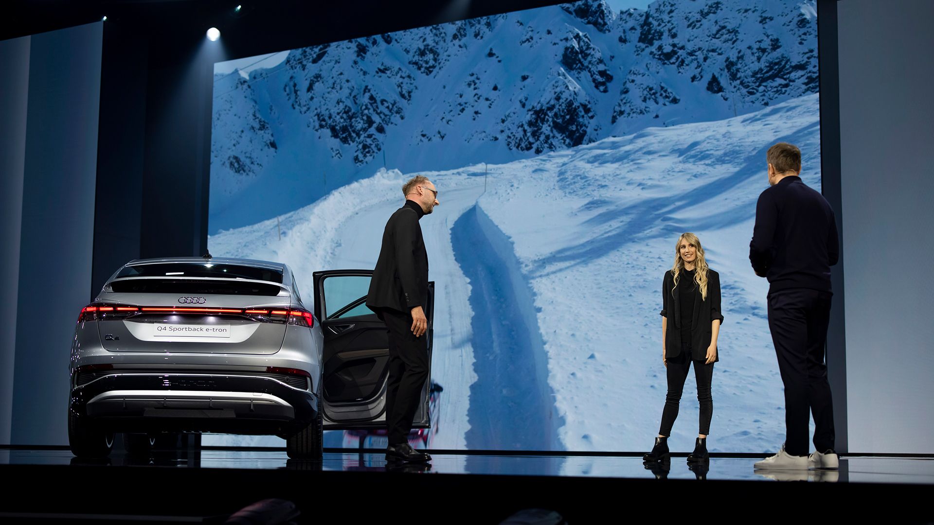 Anna Gasser stands on the stage with Marc Lichte and Steven Gätjen. A snow-covered landscape can be seen on the screen in the background.
