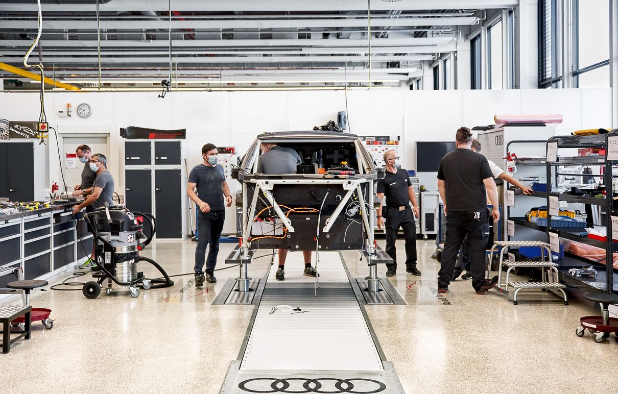 Mitarbeiter von Audi Sport arbeiten in einer Werkshalle an dem aufgebockten Käfig des Rallyeautos. 