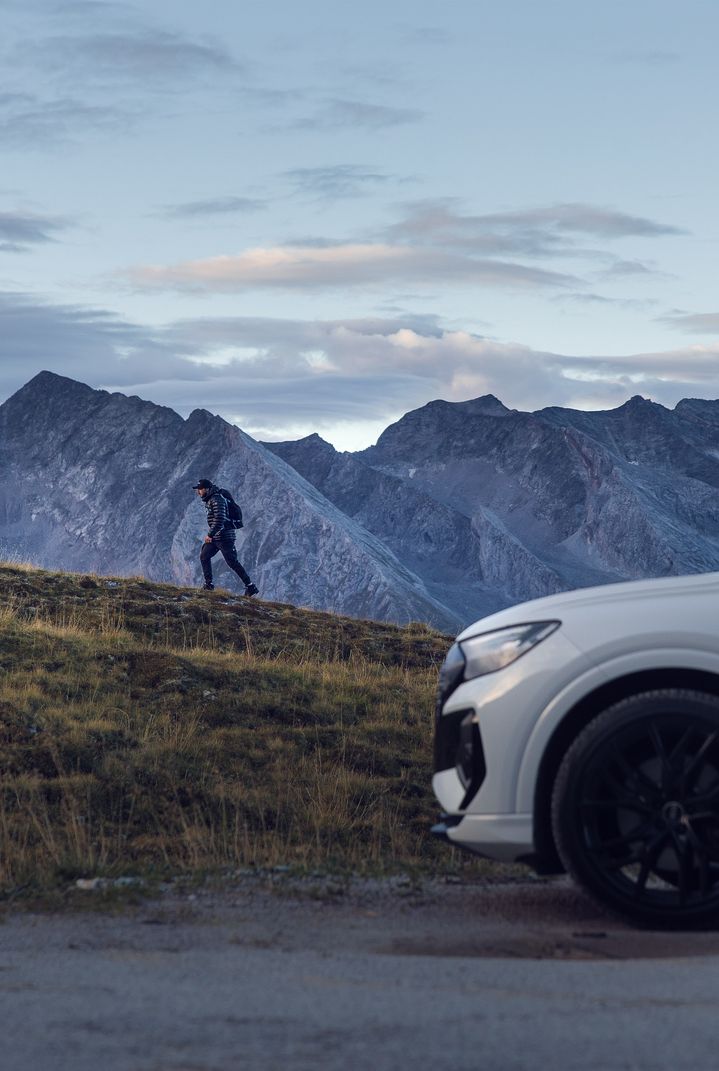 Beyaz Audi Q4 e-tron, Tom Klocker'in yürüyerek tırmandığı bir dağın önünde duruyor.