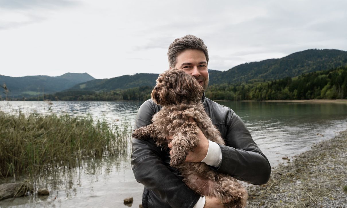 Alt-Text: Nils Wollny steht mit dem Hund auf dem Arm am See.